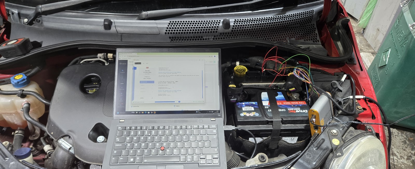 Diagnostic laptop plugged into a car's engine bay reading fault codes from the battery and ECU